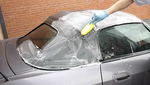 Image of a person's hand wearing a glove holding a brush and cleaning the top of a convertible grey car.